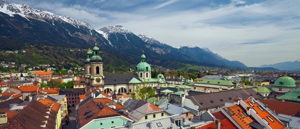 Innsbruck cityscape with baroque churches, terracotta roofs, and snow-capped Alps under a blue sky.