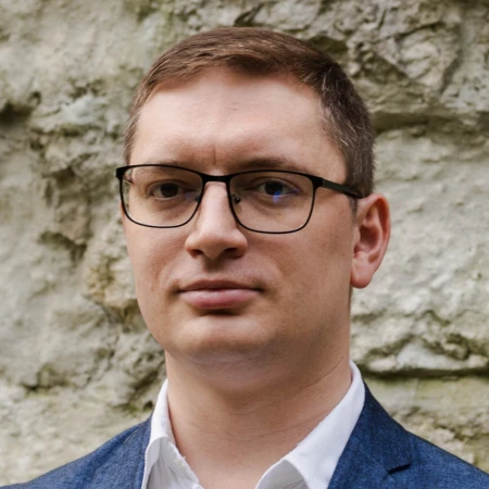 A man wearing glasses and a blue blazer, with short brown hair, looks at the camera. A stone wall is in the background.
