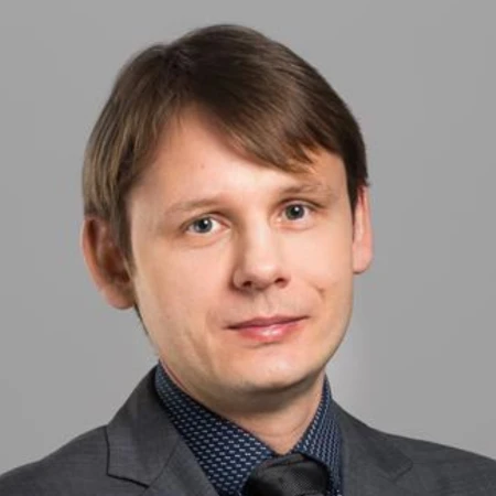 A man with light brown hair, wearing a grey suit, blue patterned shirt, and dark tie, smiles at the camera. Plain grey background.
