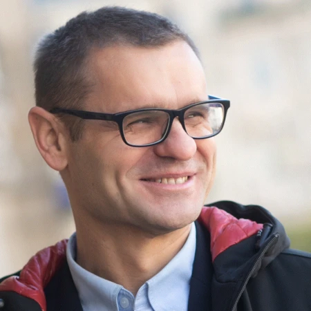 A man with short hair and black-rimmed glasses smiles warmly. He wears a light blue shirt and a dark jacket with red lining. Outdoor background.