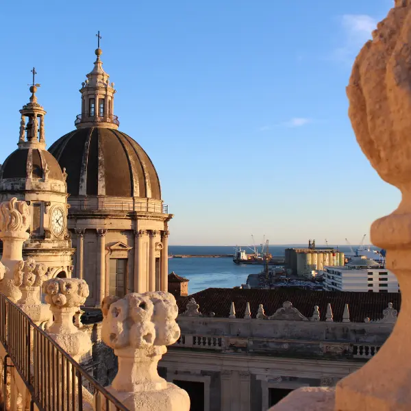 View from Abbey of St Agatha, Catania, showing domes, clock tower, ornate balustrades, port with ships, and the sea under a blue sky.