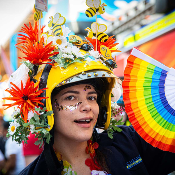 Person wears glitter on their face, wearing a highly decorated helmet and waving a rainbow fan