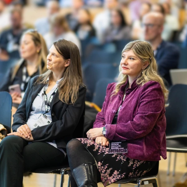 ECPR Executive Committee Members Daniela Irrera in a purple jacket and Amy Verdun in a black blazer, seated in a conference hall audience. Both wear name badges.
