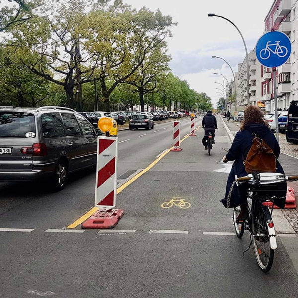 A car driving down the road. Two people are riding bikes in the cycle lane next to the road.