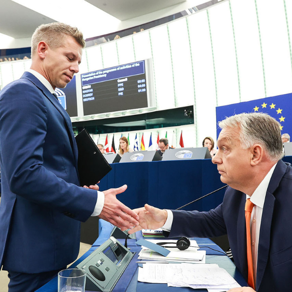 Péter Magyar is standing wearing a dark suit. He shakes hands with Viktor Orban, who is sat down wearing a dark suit. 