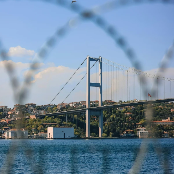 Bosphorus Bridge in Istanbul spans the blue water under a bright, cloudy sky. Buildings and green hills line the shore, partially obscured by a blurred foreground.