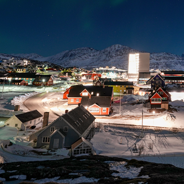 A snow-covered settlement at night with colourful houses and the aurora borealis visible in the starry sky. Mountains loom in the background.