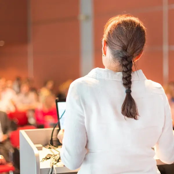 Woman with braided hair, facing away from camera, presenting at a podium in front of an audience.
