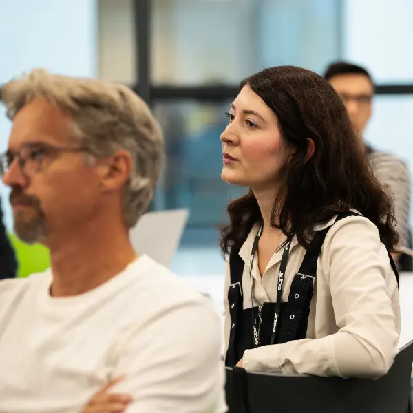 A woman and a man seated a classroom, both attentive. The woman has dark hair and is wearing a white shirt with black straps.