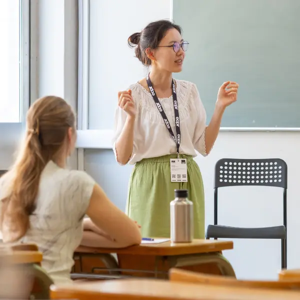 Woman presenter in glasses, white blouse, and green skirt gestures while speaking at the ECPR conference. Member of the audience in foreground.