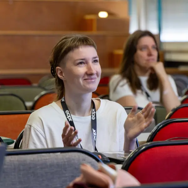Woman with short fringe and undercut hairstyle smiles and claps, wearing a white t-shirt and ECPR lanyard. Lecture hall setting.