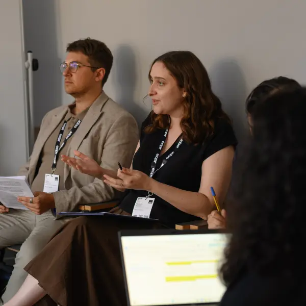 A group of people wearing ECPR lanyards seated during a panel at the ECPR conference. A woman in a black short sleeved top speaks animatedly, a man in a beige jacket listens, and a laptop is visible.