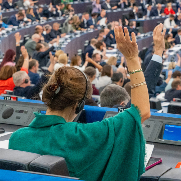 A close up woman sat in the European Parliament raises her hand, wearing a green scarf. Many other people are also raising their hands in the background.