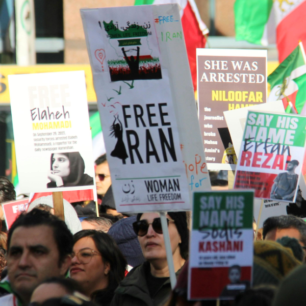 People in a crowd hold signs and Iranian flags. The largest sign says 