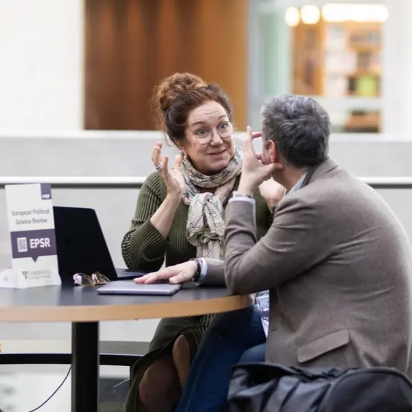 Woman gesturing and animatedly talking to a man at a table with a laptop and a sign for European Political Science Review.