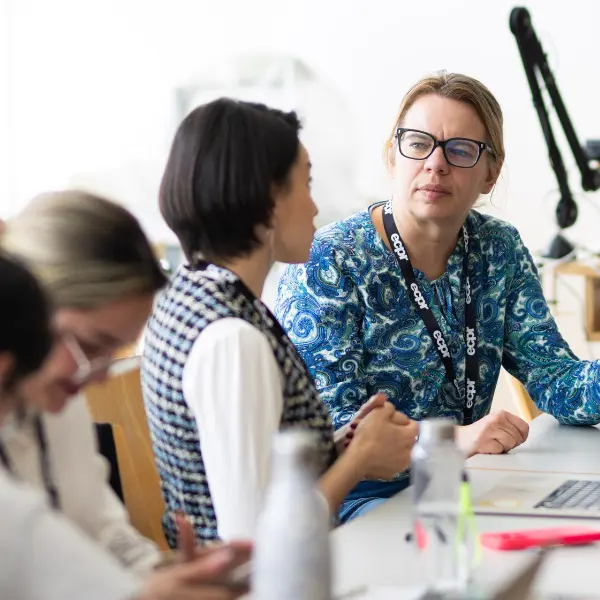 Three people in a discussion at a table during an ECPR workshop. One woman wears glasses and a blue patterned top with an ecpr lanyard. Another wears a checked vest. A third looks down.