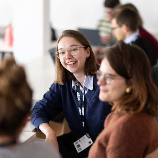 Two young women smiling and talking. The woman on the left wears glasses and a navy jumper with an ECPR lanyard. The woman on the right wears glasses and a rust jumper.