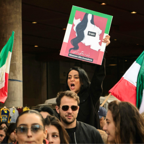 A crowd of people gather, some are holding Iran flags. One woman carries a sign which reads Iran, Women Life Freedom.