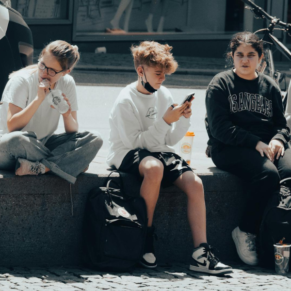 Three teenagers sit on a wall wearing monochrome outfits. One looks at a phone, one is drinking from a Starbucks cup, the other looks bored and straight ahead.