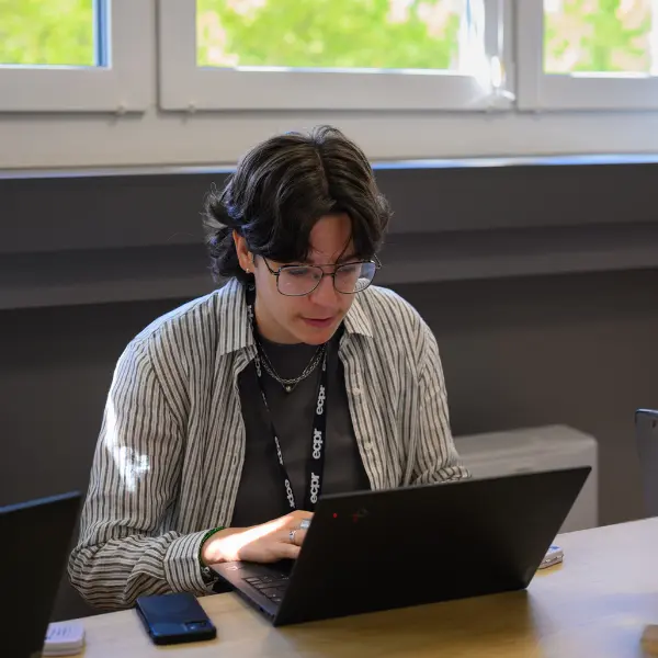 Young person, wearing glasses and striped shirt, seated in a classroom setting in front of a window, working on a laptop. The person wears an ecpr lanyard. Smartphone visible on the table.