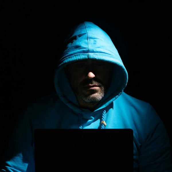 Man in a bright blue hoodie with his face in shadow, looking down at a laptop screen in a dark room.