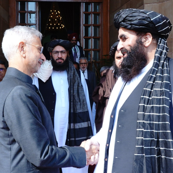 India External Affairs Minister Dr. S. Jaishankar and Minister of Foreign Affairs of Afghanistan, Amir Khan Muttaqi shake hands outside a building. Jaishankar wears a grey suit, Muttaqi a black turban and waistcoat over a white shirt with striped scarf.