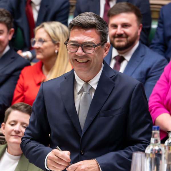 Andy Burnham is smiling in the Houses of Parliament wearing a dark suit and grey tie. 