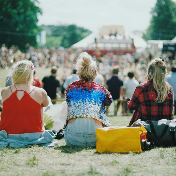 Three women at a festival sit on the grass facing away from the camera