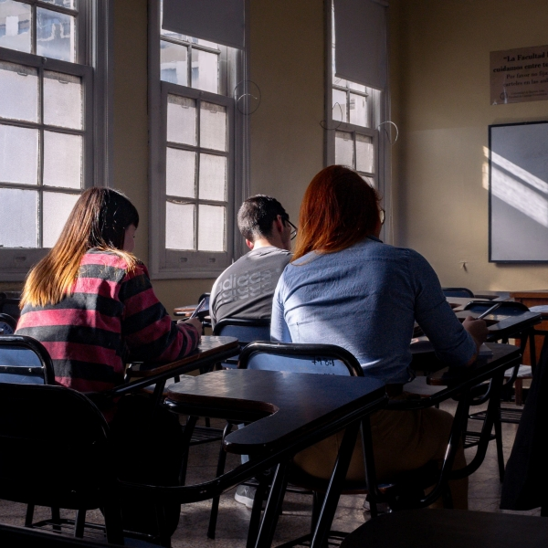 Three people seated in a classroom, taking notes, with ambient light coming from large windows on the left.