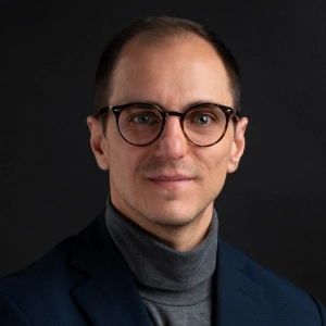 Headshot of Andrea De Angleis against a dark background. He wears tortoiseshell glasses and a navy blue blazer over a grey roll-neck jumper.