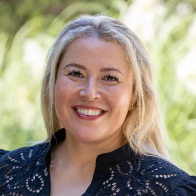 Headshot of Tanja Munro, a smiling woman with blonde hair, wearing a dark patterned top. Outdoors with blurred green foliage.